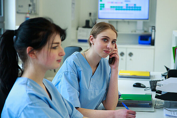 Zwei medizinische Fachkräfte auf der Station. Eine Fachkraft telefoniert.