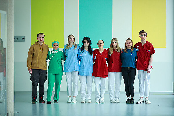 Pflegeteam vor einer bunten Wand in der Klinik haben sich im Arm. Sie tragen unterschiedliche Arbeitskleidung